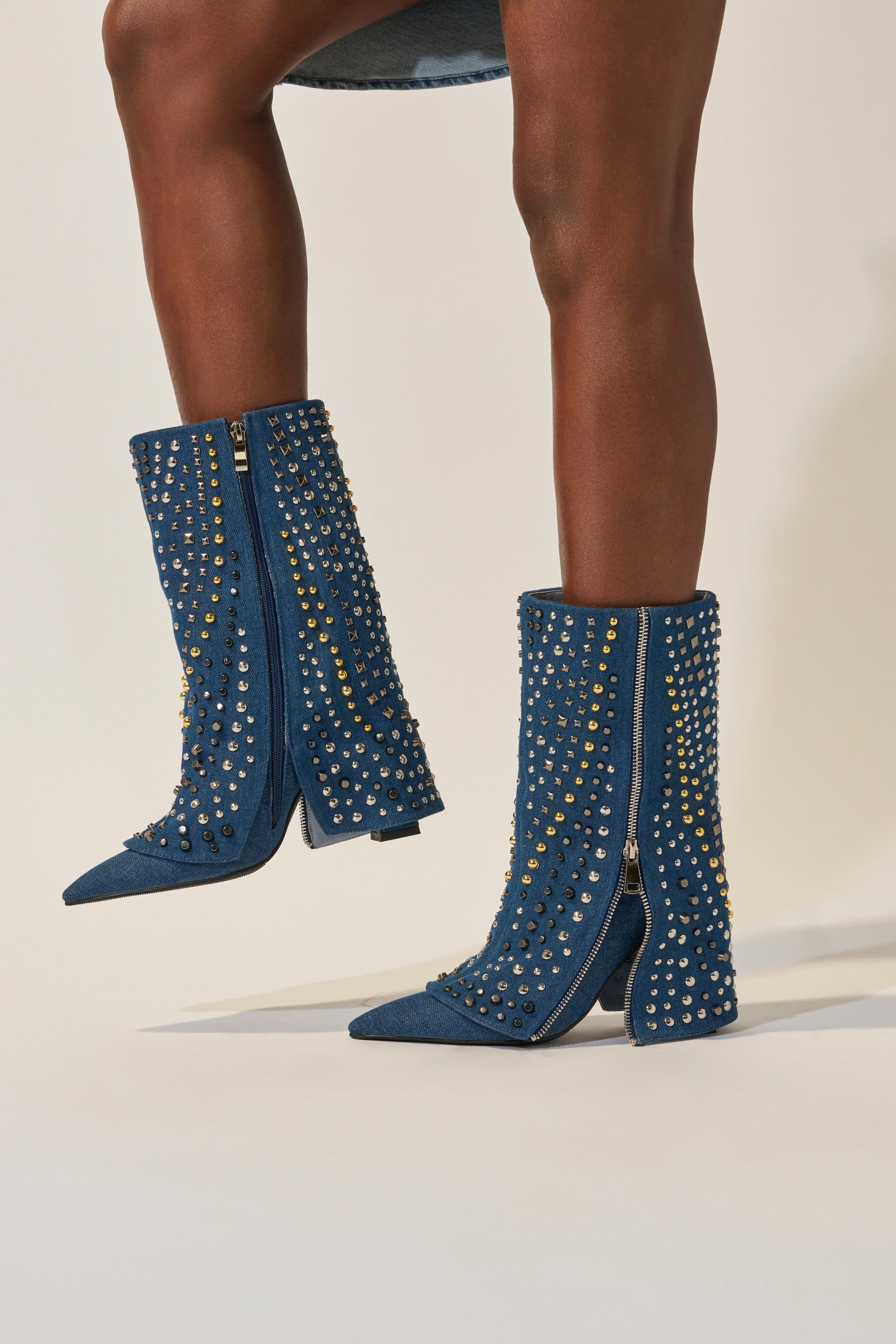 Someone wears the AZALEA WANG HENZI DENIM STUDDED CHUNKY BOOTIE—blue denim with pointed toes, gold and silver studs, side zippers, and chunky heels—styled with a short denim skirt against a neutral background. 2