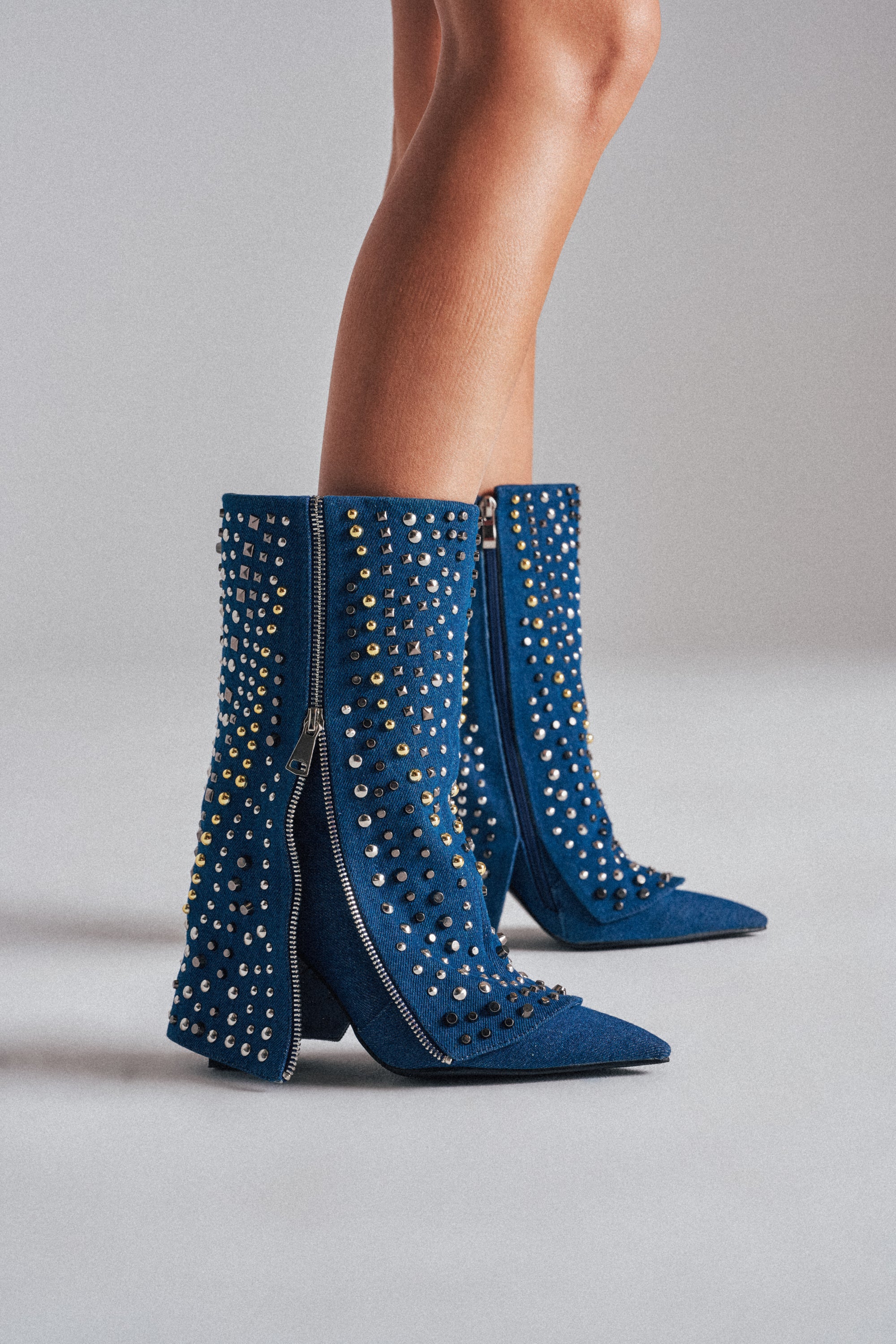 A person wears the AZALEA WANG HENZI DENIM STUDDED CHUNKY BOOTIE, featuring mid-calf blue denim, silver and gold studs, pointed toes, chunky heels, and side zippers on a light gray surface. 0