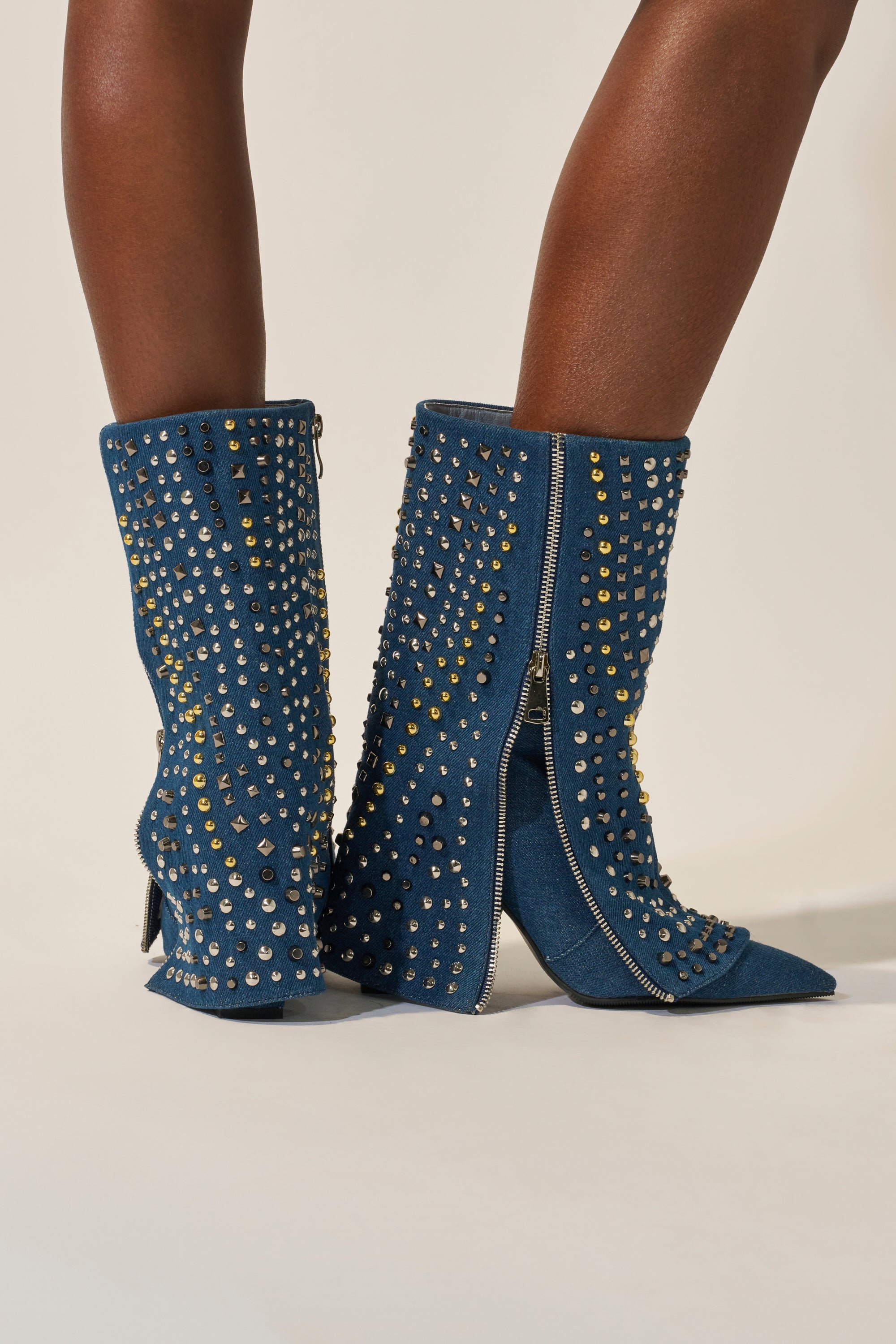 A person wears the AZALEA WANG HENZI DENIM STUDDED CHUNKY BOOTIE, a mid-calf denim boot with gold and silver studs, side zippers, pointed toes, and a chunky heel, standing on a light background. 3