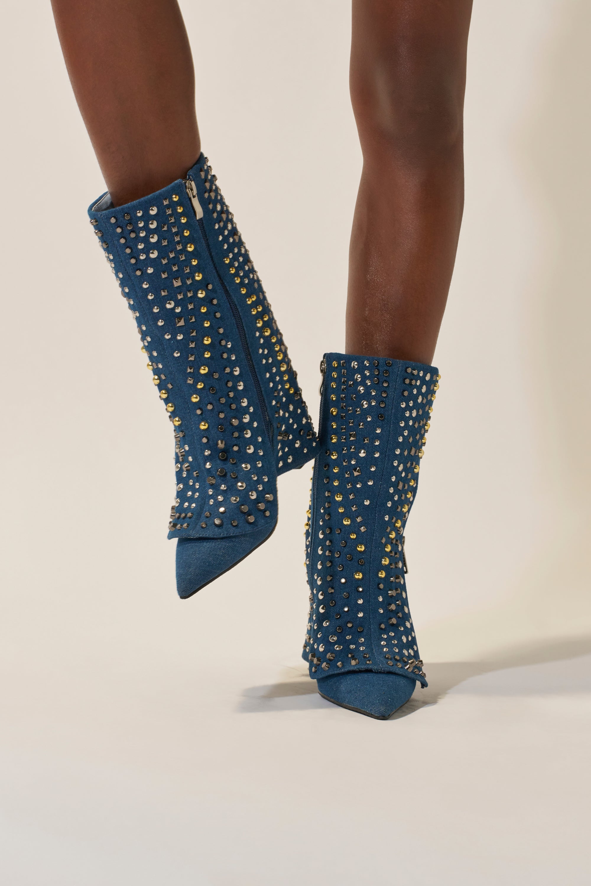 A person stands on a light-colored surface wearing the AZALEA WANG HENZI DENIM STUDDED CHUNKY BOOTIE—blue pointed toe booties with silver and gold studs and a side zipper. 4