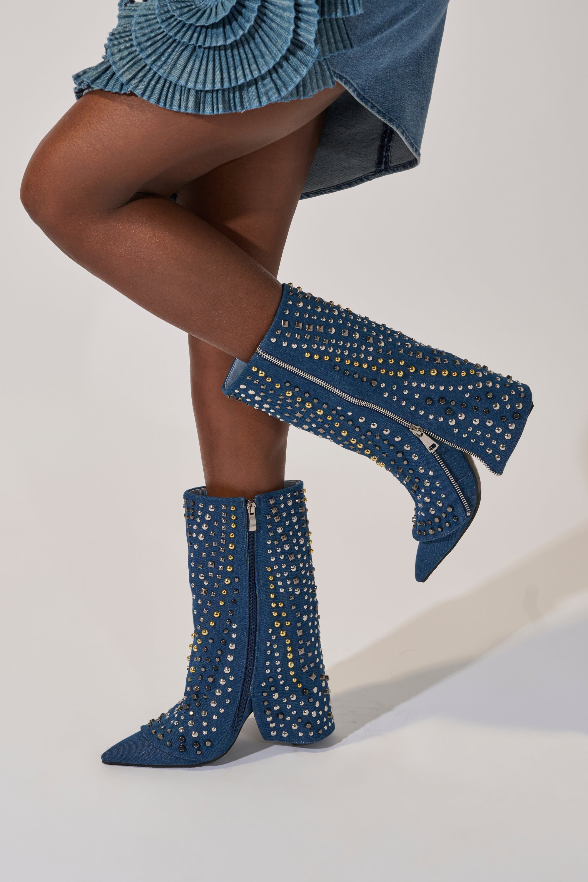 A person stands with legs crossed against a plain background, wearing a blue denim skirt and the AZALEA WANG HENZI DENIM STUDDED CHUNKY BOOTIE—pointed toe denim boots adorned with silver and gold studs. 9