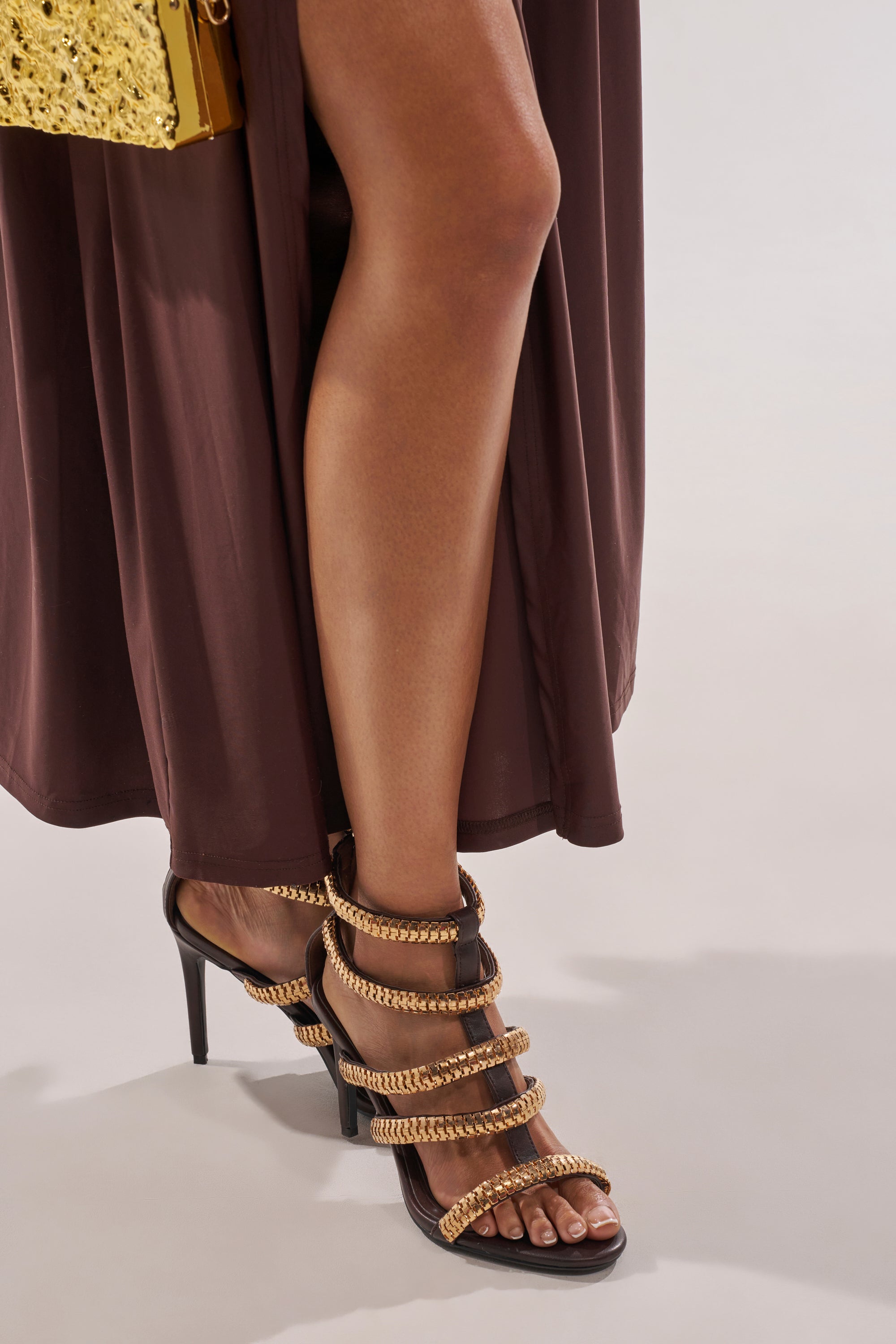 Wearing a brown dress and AZALEA WANG HERSILIE BROWN GLADIATOR STILETTO heels with gold chain accents, and holding a gold textured clutch, a person stands on a light floor. 1