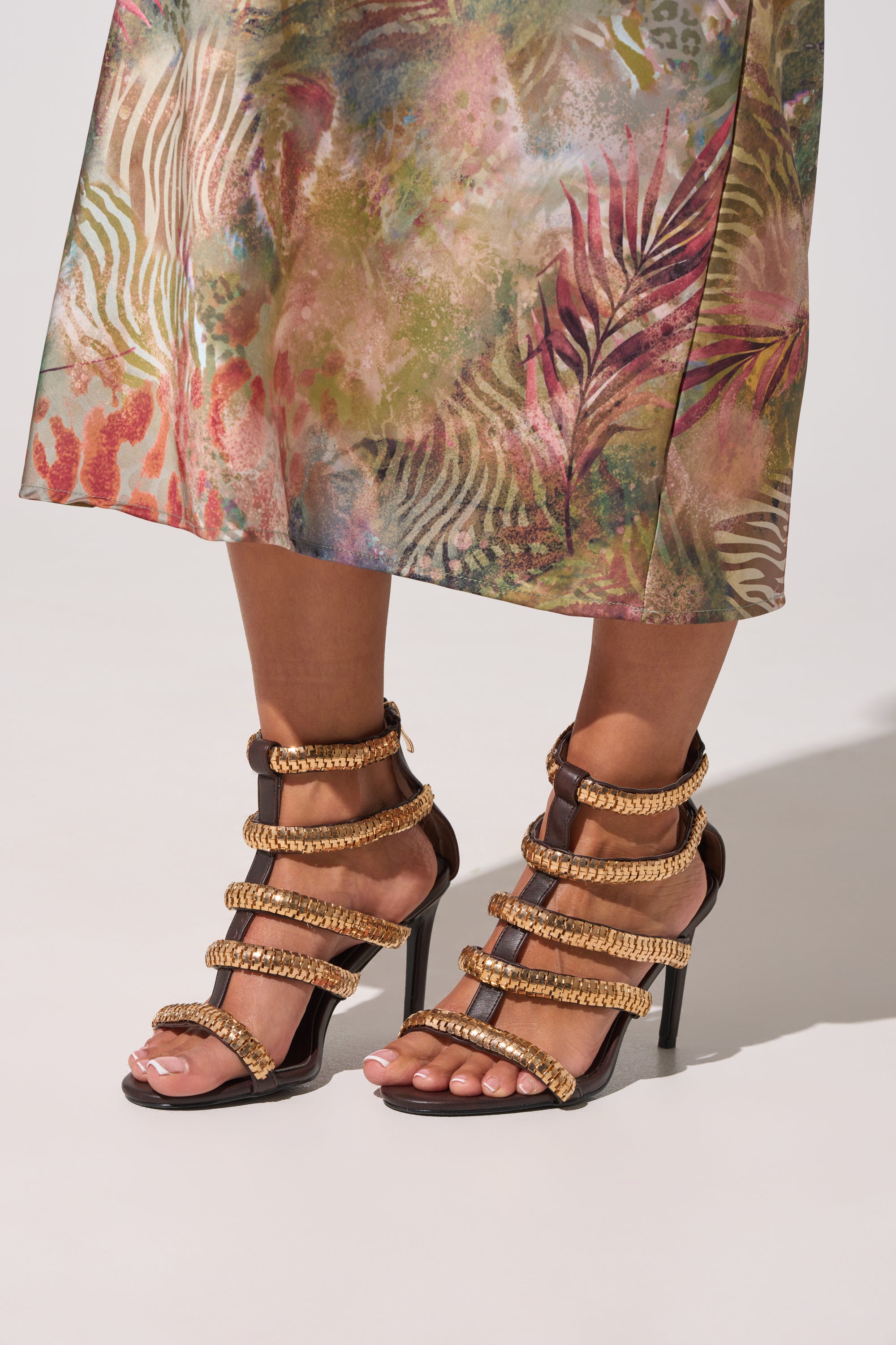 A person stands in AZALEA WANG HERSILIE BROWN GLADIATOR STILETTO sandals, featuring tan braided straps, paired with a colorful tropical skirt and painted toenails, set against a light background. 0