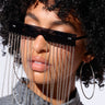 A person with curly hair is wearing CHAIN TEARS SUNGLASSES, which feature silver chain link accents hanging from the lenses. They are also adorned with large hoop earrings and a gray denim shirt. The plain white background perfectly highlights these unique fashion accessories.