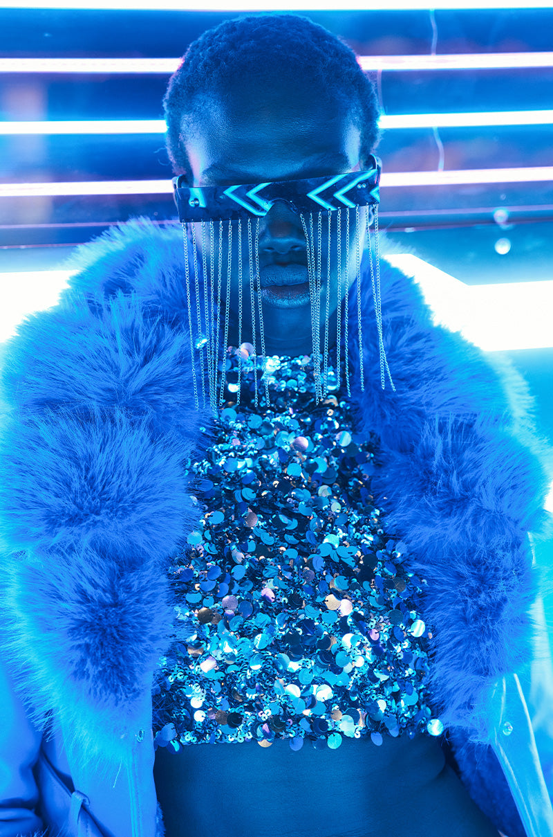 A person is wearing CHAIN TEARS SUNGLASSES, characterized by their futuristic, angular design with dangling elements, along with a fluffy blue coat. Their sequined top captures the vivid neon-lit environment, crafting a vibrant color scheme highlighted by shades of blue.
