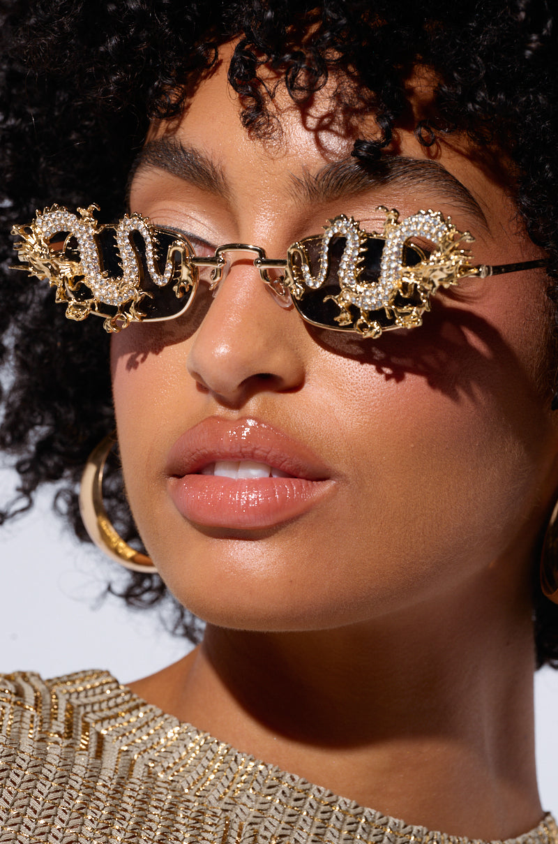 Close-up of a person with curly hair wearing DRAGON EYES SUNGLASSES, shaped like snakes and featuring intricate, rhinestone-adorned dragon detailing. The sunglasses have a faux gold metallic frame. The person is also wearing gold hoop earrings and a textured gold top. Their expression is neutral against a plain white background.
