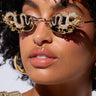 Close-up of a person with curly hair wearing DRAGON EYES SUNGLASSES, shaped like snakes and featuring intricate, rhinestone-adorned dragon detailing. The sunglasses have a faux gold metallic frame. The person is also wearing gold hoop earrings and a textured gold top. Their expression is neutral against a plain white background.