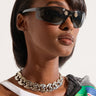 A person with short dark hair is wearing PIERCING SUNGLASSES, a white tank top, a chunky silver chain necklace, and a jacket with a colorful print draped over one shoulder. The background is white.