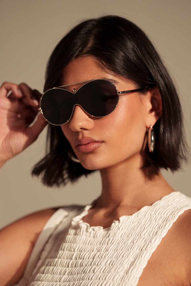 A woman with a sleek bob wears large, black MEME SUNGLASSES and gold hoops. Dressed in a textured white sleeveless top, she poses against a neutral background, touching her sunglasses with one hand.