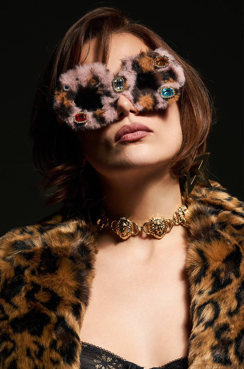 A person with short hair wears oversized AMORE FUR GLASSES featuring gemstone accents and a leopard print design. They complement this look with a matching leopard print coat and a bold gold necklace, all set against a dark background.