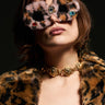 A person with short hair wears oversized AMORE FUR GLASSES featuring gemstone accents and a leopard print design. They complement this look with a matching leopard print coat and a bold gold necklace, all set against a dark background.