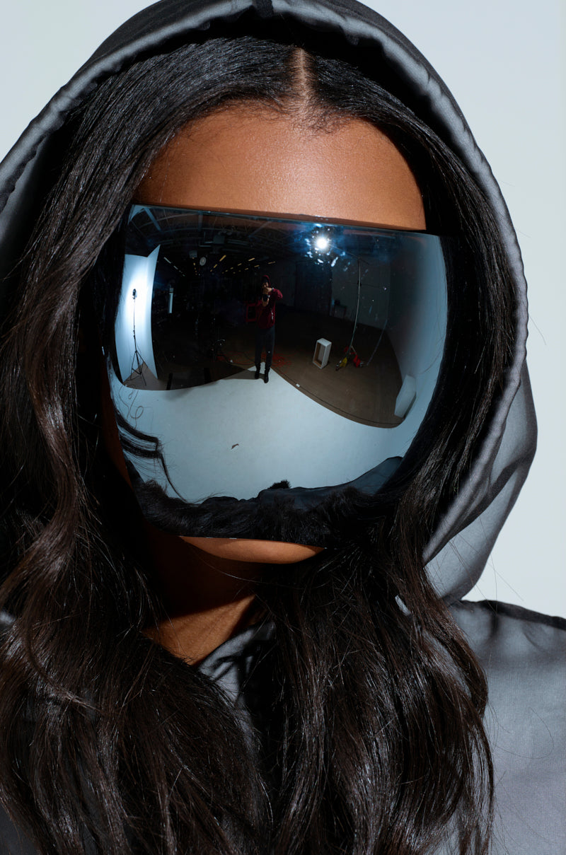 A person wears the PROTECT YA SELF FACE SHIELD with a large mirrored visor and dark hood. The visor reflects a room with lights and equipment. Long hair cascades over their shoulders, contrasting against the minimal, bright background. 1