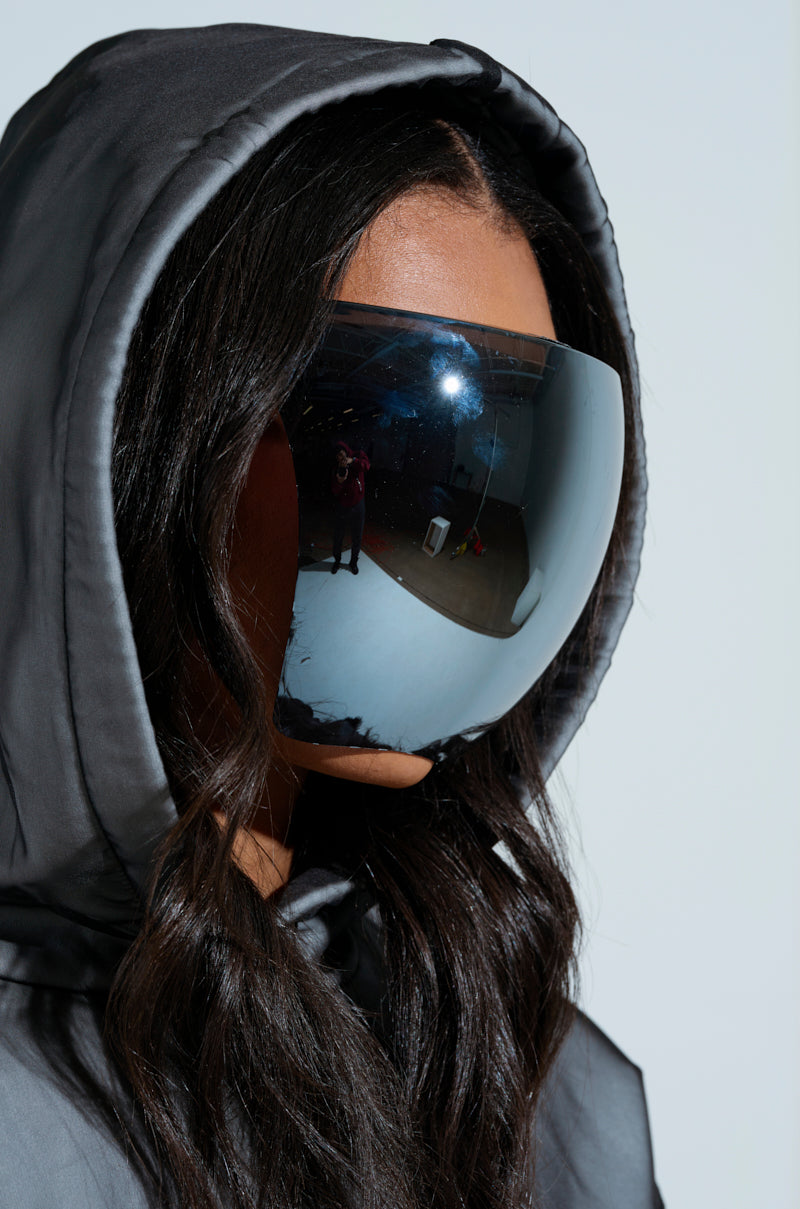 A person dons the PROTECT YA SELF FACE SHIELD, a reflective mirrored visor akin to oversized sunglasses, paired with a dark hooded jacket. The shield reflects the photographer and surroundings, creating a futuristic and mysterious appearance. 2