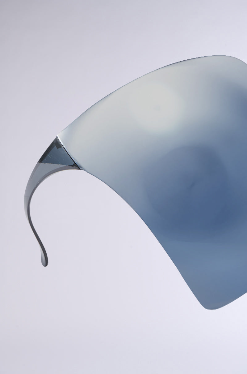 The PROTECT YA SELF FACE SHIELD features a sleek, futuristic design with a reflective, curved surface in light blue and grey tones against a plain background, blending technology and style into its smooth, minimalist look. 4
