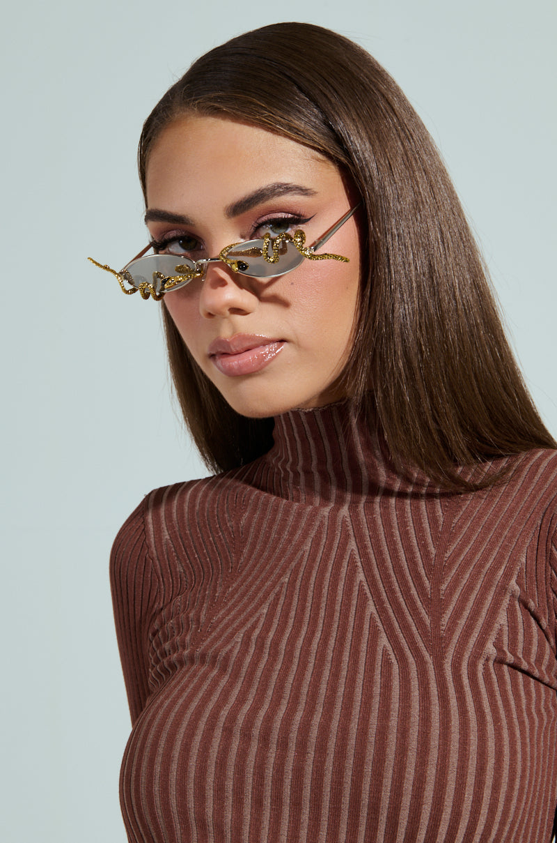 A person with long brown hair wears DONT BE A SNAKE SUNGLASSES featuring reflective oval lenses and rope-like detailing. They are dressed in a ribbed, long-sleeve brown turtleneck, against a light neutral background, creating a minimalist look.
