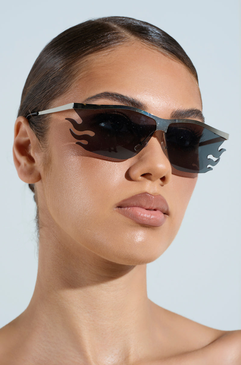A person with sleek dark hair and bold eyebrows wears FEEL THE FLAME SUNGLASSES with rectangular tinted lenses. The plain background accentuates the unique eyewear and the person's confident expression, while adjustable nose pads ensure comfort.