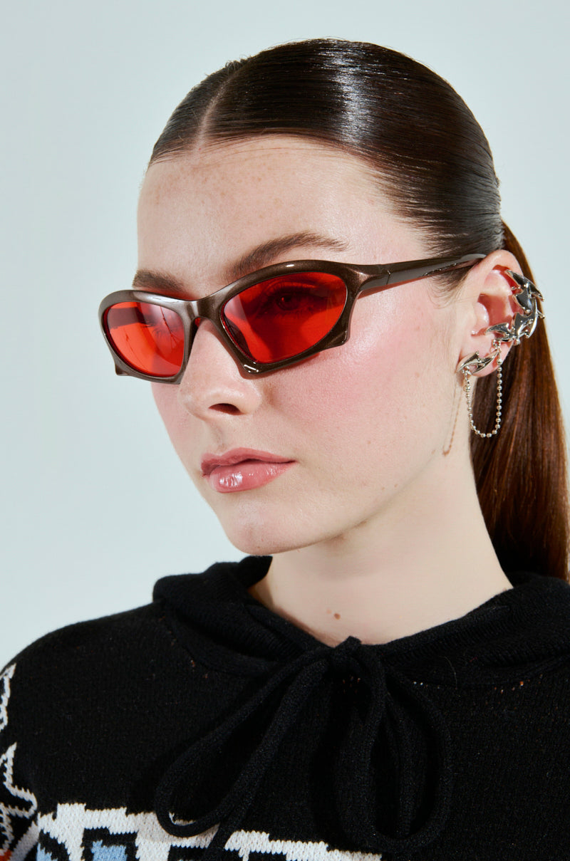 A person with straight, dark hair pulled back wears SEEING RED SUNGLASSES with a brown plastic frame and a black top with a white pattern. They sport an intricate ear cuff adorned with chains against a plain, light-colored background. 1