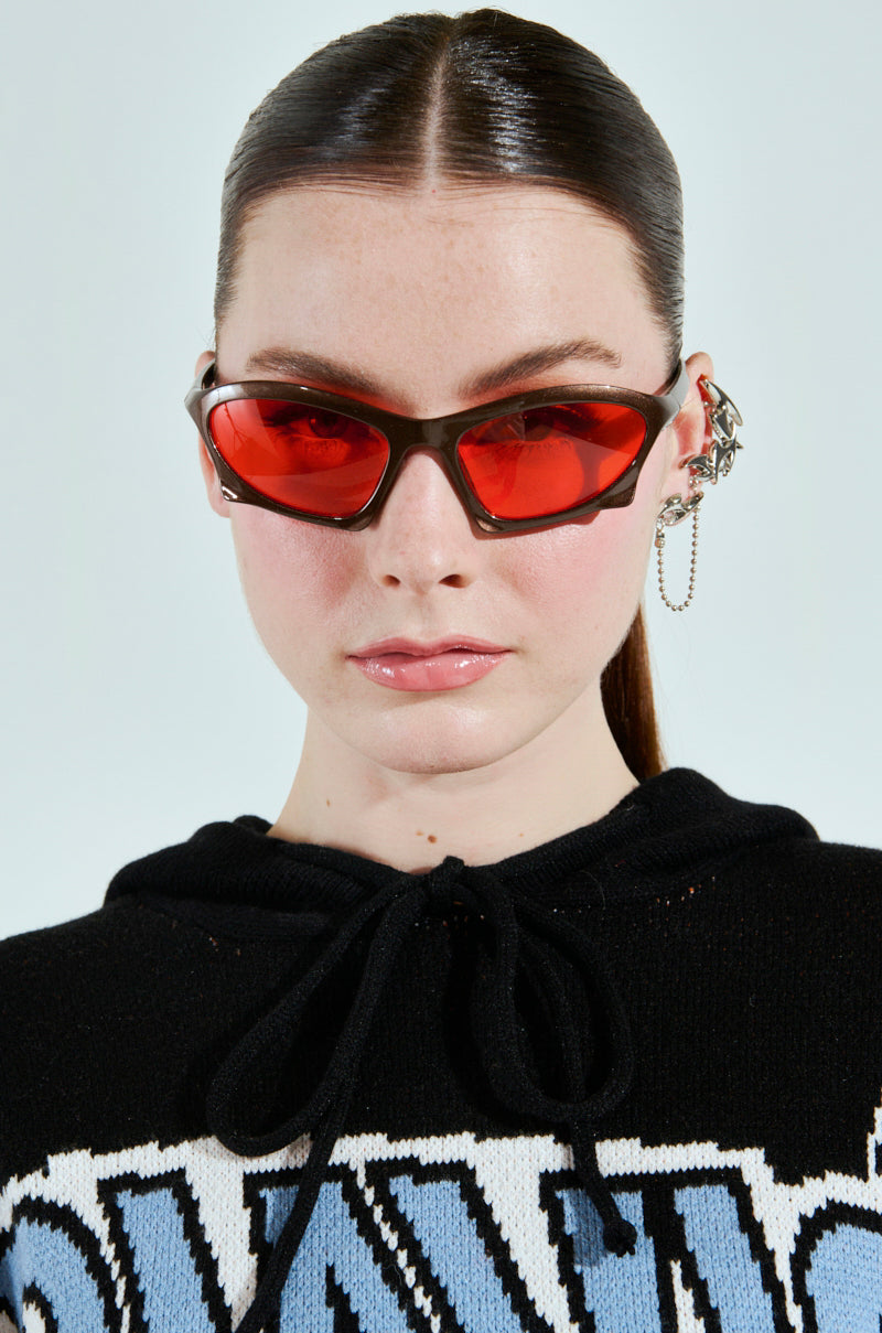 A person with long hair tied back wears SEEING RED SUNGLASSES with brown plastic frames and an ornate chain ear cuff. They have a neutral expression, dressed in a patterned black and white top, against a plain background. 2