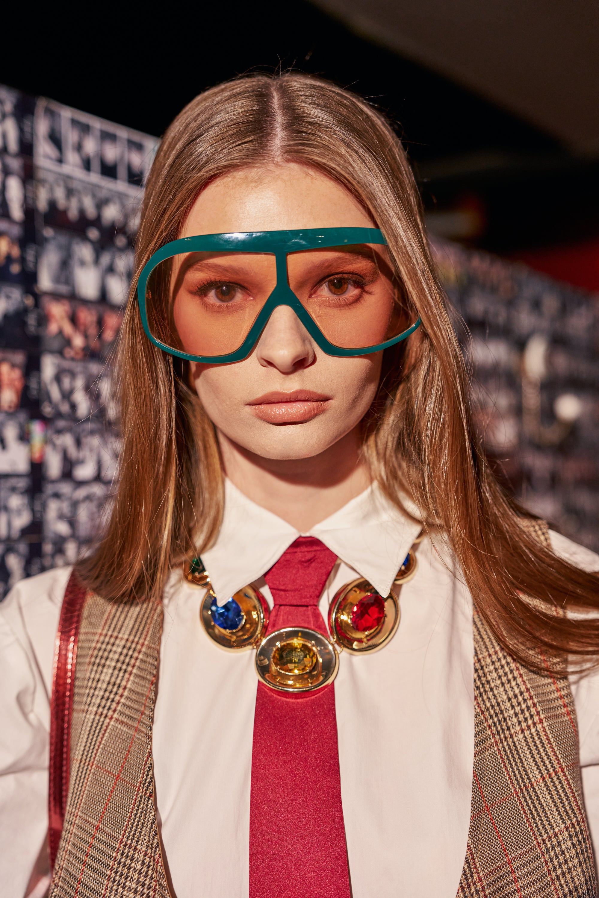A woman wearing oversized green shield ALYSSA SUNGLASSES, a white collared shirt, a red tie, and a statement necklace with large, colorful circular gems.