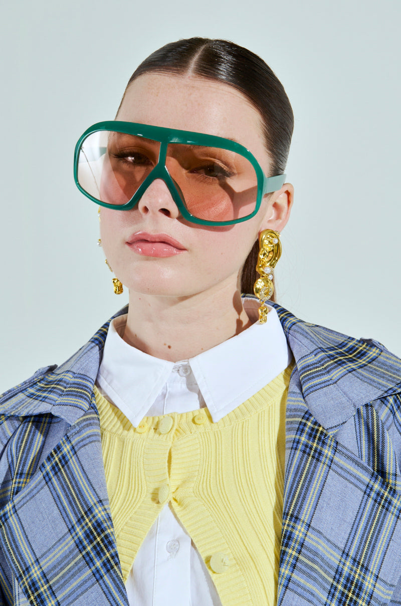 Wearing oversized green ALYSSA SUNGLASSES and gold earrings, the person sports a plaid jacket over a yellow textured top and white collared shirt. With slicked-back dark hair, they have a neutral expression against a plain light gray background, adding to their cool, understated vibe. 3