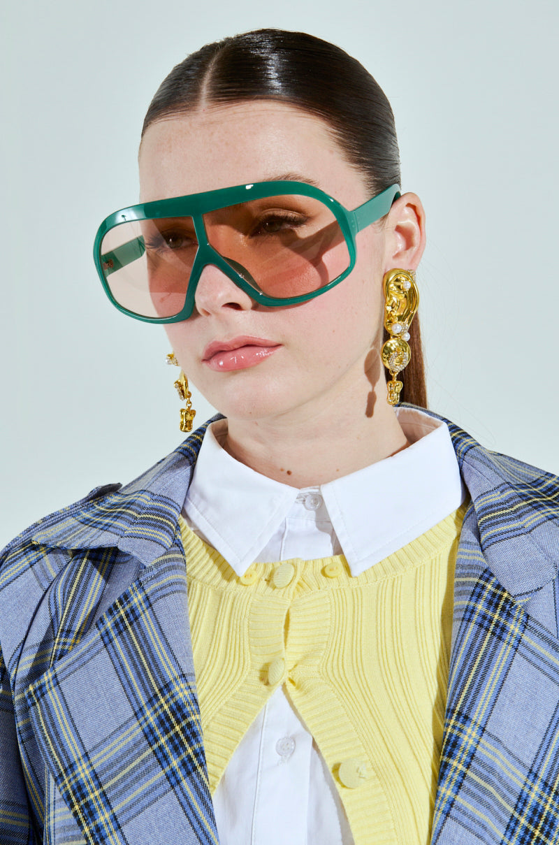 A woman with long dark hair wears green ALYSSA sunglasses featuring oversized lenses, gold statement earrings, a yellow sweater over a white collared shirt, and a plaid blazer. She gazes forward with a neutral expression against a plain background. 4