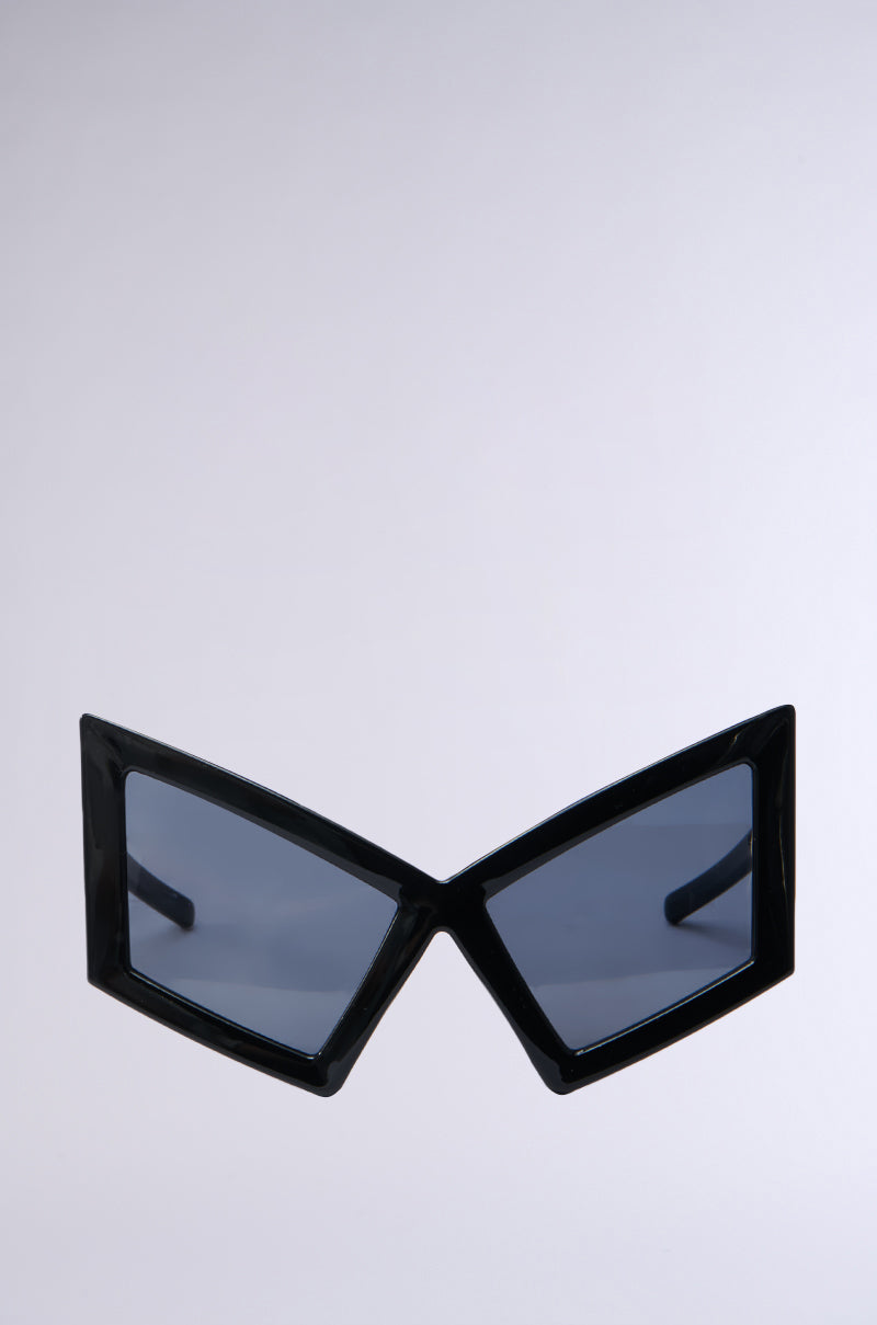 The U HAVE TO GET IT SUNGLASSES offer a Matrix-chic vibe with black futuristic geometric frames and oversized temple arms. Tinted lenses add mystery, set against a plain, neutral background.