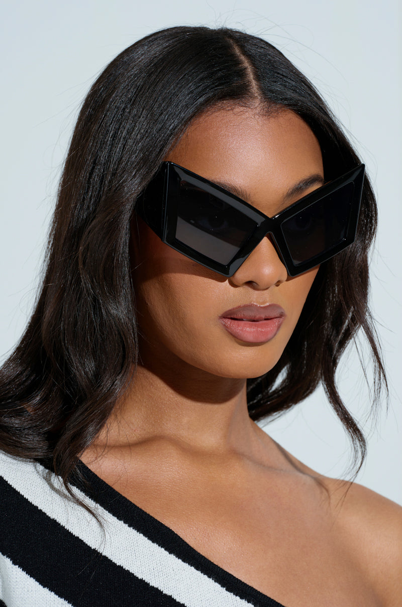 A woman with long dark hair models the Matrix-chic "U HAVE TO GET IT" sunglasses, showcasing their large geometric black frames and oversized temple arms. Her one-shoulder black and white striped top stands out against the plain white background.