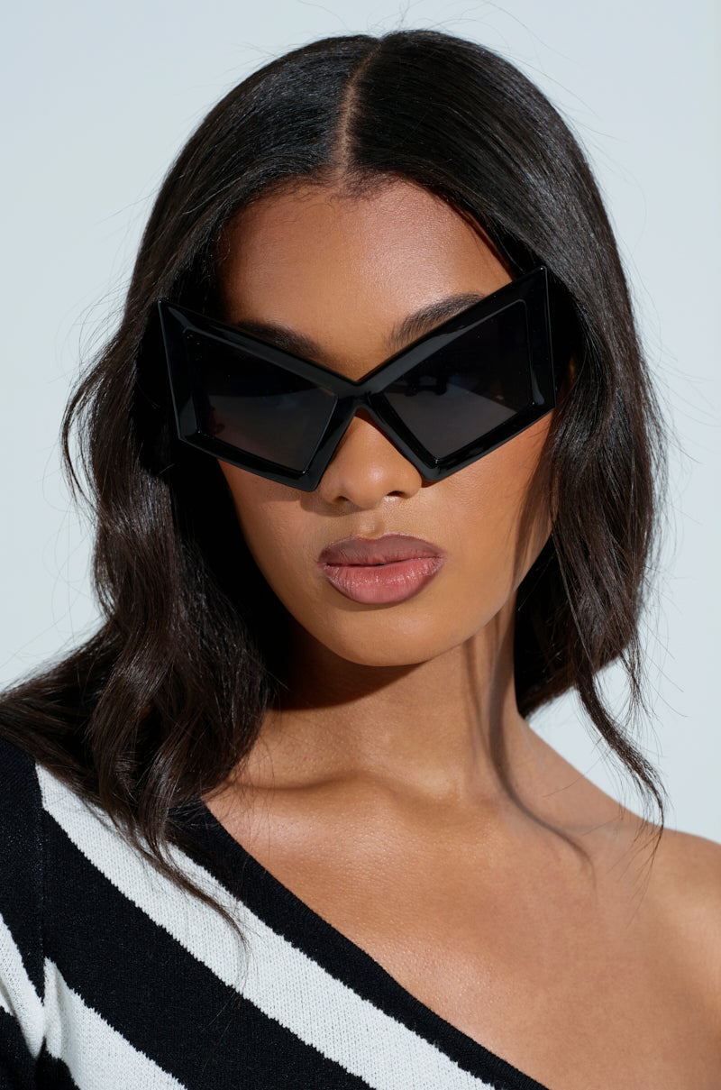 A woman with long black hair sports the U HAVE TO GET IT SUNGLASSES, featuring Matrix-chic oversized temple arms and tinted lenses. She pairs them with a black-and-white striped top with an asymmetrical neckline, set against a solid light gray backdrop.