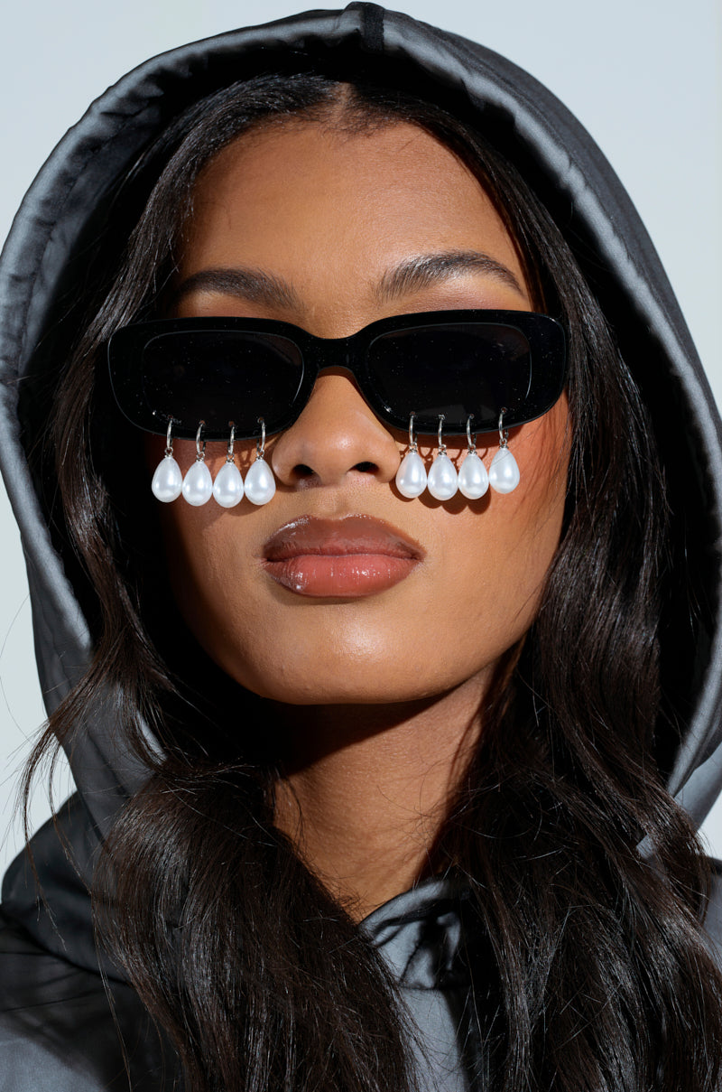 A person in a gray hooded jacket wears PEARL PLZ SUNGLASSES, featuring black frames with tinted rectangular lenses and teardrop pearl embellishments that hang elegantly. Their long hair flows over their shoulders.