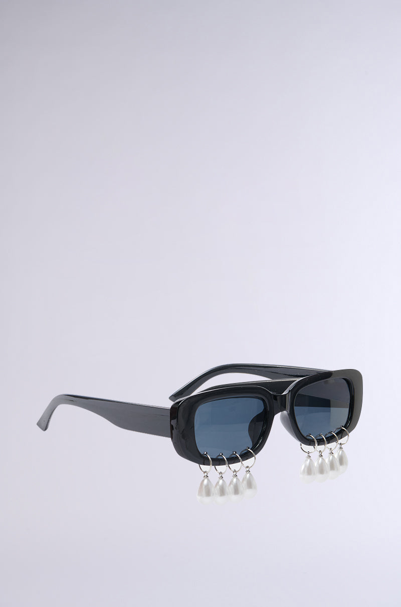 Introducing the PEARL PLZ SUNGLASSES: sleek black frames with tinted rectangular lenses and distinctive teardrop pearl embellishments, all set against a plain gray backdrop.
