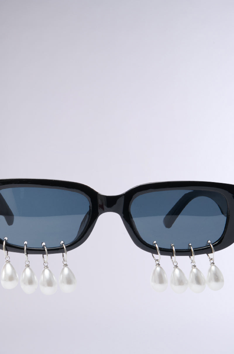 Introducing the elegant PEARL PLZ SUNGLASSES: black rectangular frames with unique blue-tinted lenses, enhanced by teardrop pearl embellishments. With a simple light gray backdrop, these sunglasses promise to elevate any look with sophistication and style.