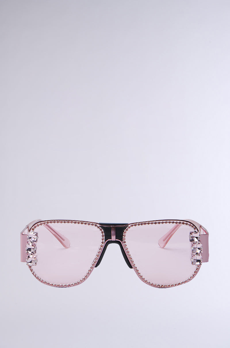 The PRETTY IN PINK SUNGLASSES are oversized aviator-style with a black front bridge and pink lenses. They feature rhinestone trim along the edges and sparkling crystal embellishments on the sides, set against a plain light gray background.