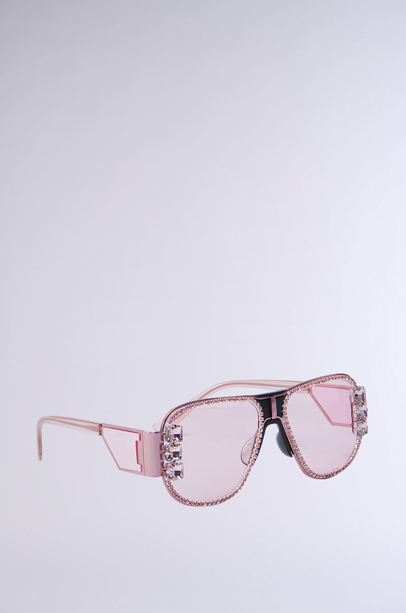 PRETTY IN PINK SUNGLASSES feature large rectangular pink lenses with rhinestone trim on the top and sides, complemented by thin matching temples. Set against a plain light gray background, these shades offer a striking look.