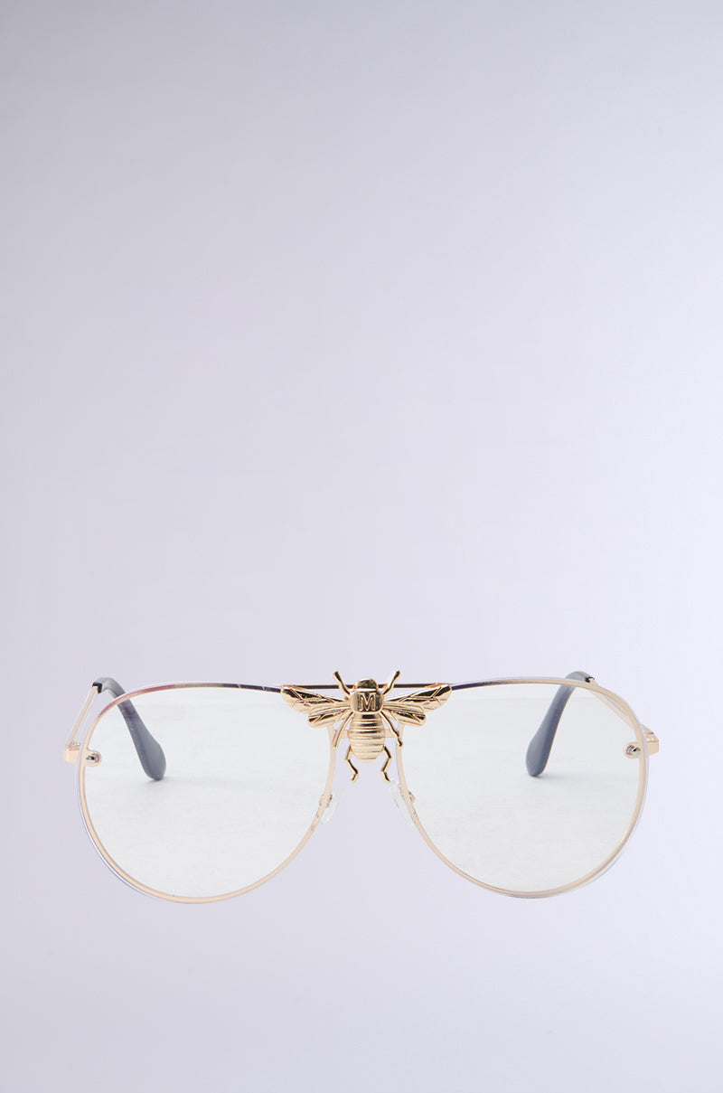 The FEEL ME BEE GLASSES by AKIRA Label showcase gold-rimmed, monobrow aviator lenses, highlighted with a decorative gold bee on the bridge. These stylish sunglasses include light-colored lenses and slender black arms set against a simple light gray backdrop. 0