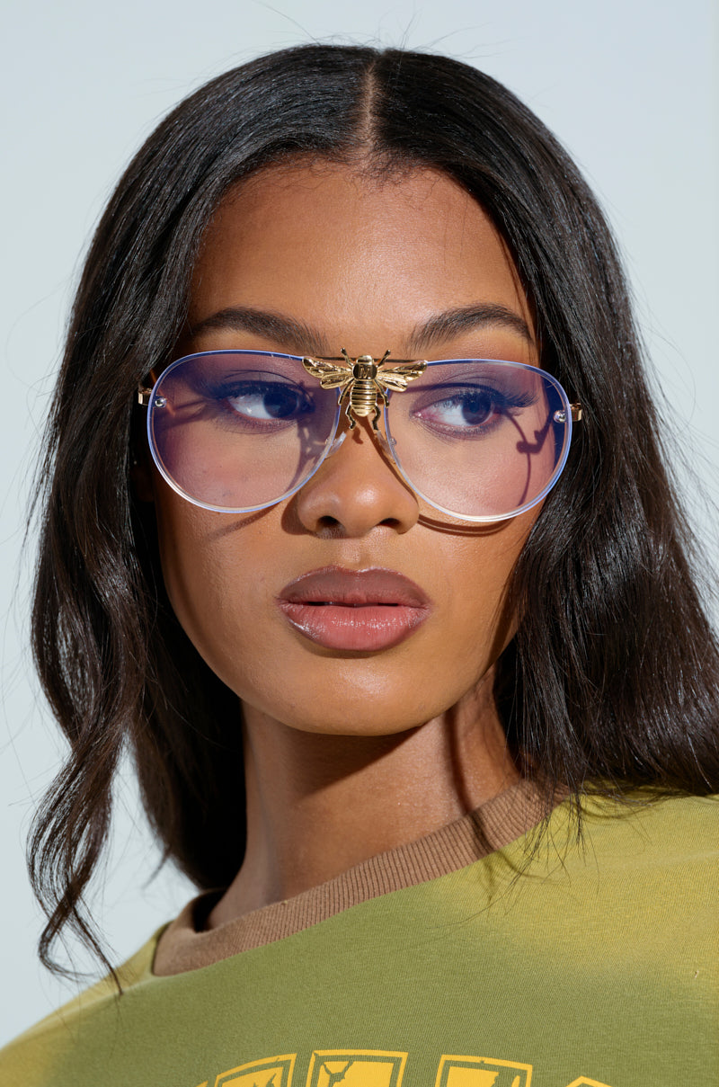 A woman with long dark hair wears large, round glasses with a gold bee detail on the bridge, resembling the FEEL ME BEE GLASSES. She has a serious expression and wears a green shirt with yellow accents against a light, blurred background. 1