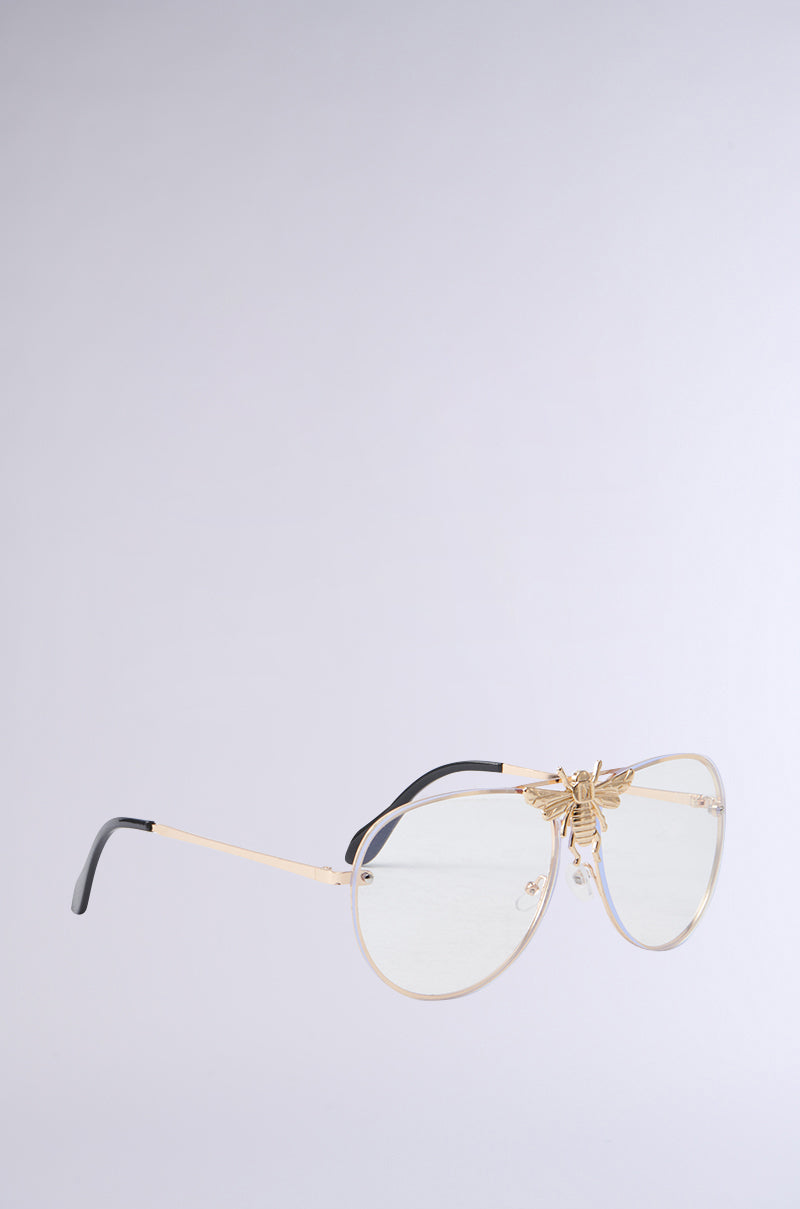 The FEEL ME BEE GLASSES feature monobrow aviator-style lenses with gold frames, translucent lenses, and a decorative gold bee on the bridge. They also have black temple tips set against a light gray background. 2