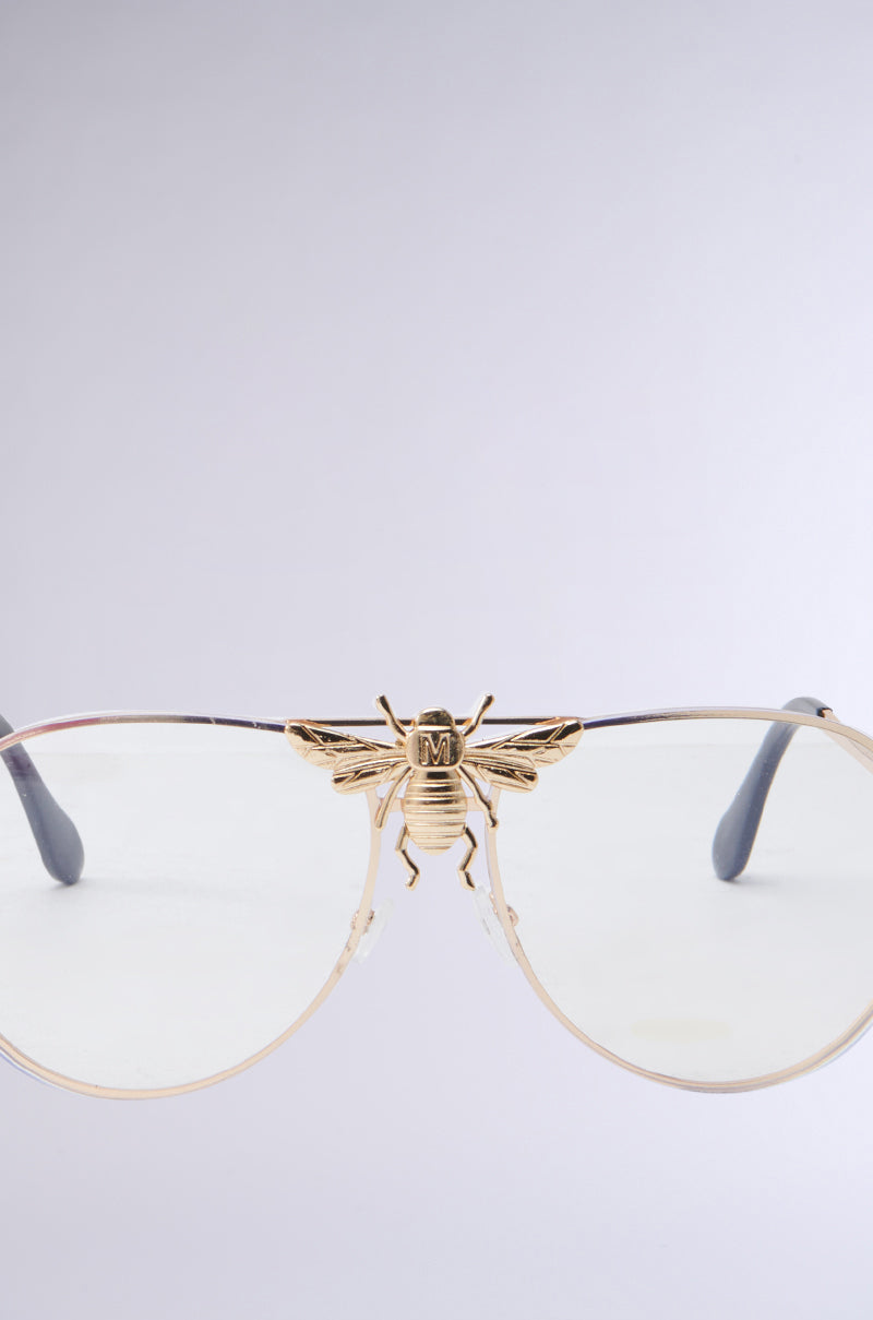 Close-up of FEEL ME BEE GLASSES, showcasing a gold bee decoration at the bridge. The monobrow aviator-style frames are silver with clear lenses, against a soft gray gradient background. 3