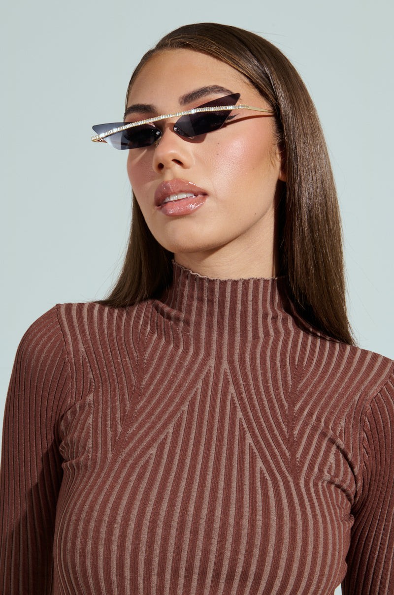 A person with long, straight brown hair is confidently posing against a light blue background, wearing GET ALIGNED SUNGLASSES featuring rhinestone embellishments and a ribbed high-neck brown top, while looking to the side. 2