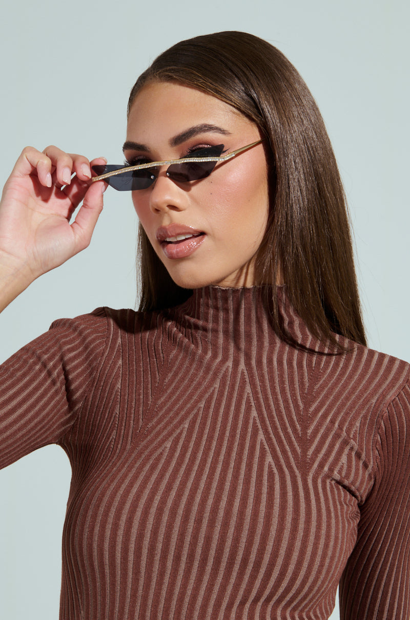 A woman with straight hair and bold eyeliner wears a brown, ribbed high-neck top. She slightly lowers GET ALIGNED SUNGLASSES on her nose while gazing forward. Her makeup features soft lipstick, set against a light, neutral background. 3