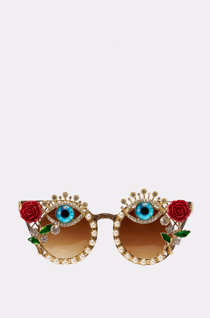 OFF THE RUNWAY SUNGLASSES with a gold frame, adorned with pearls, jeweled eyelashes, red rose accents, and blue eye designs above each lens, showcased on a plain white background. 0
