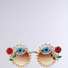 The OFF THE RUNWAY SUNGLASSES feature pearl-encrusted gold frames. Each lens, adorned with a blue eye design and rhinestones, is stylishly accented with red roses and green leaves in the corners. The brown-tinted lenses complete this elegant accessory.