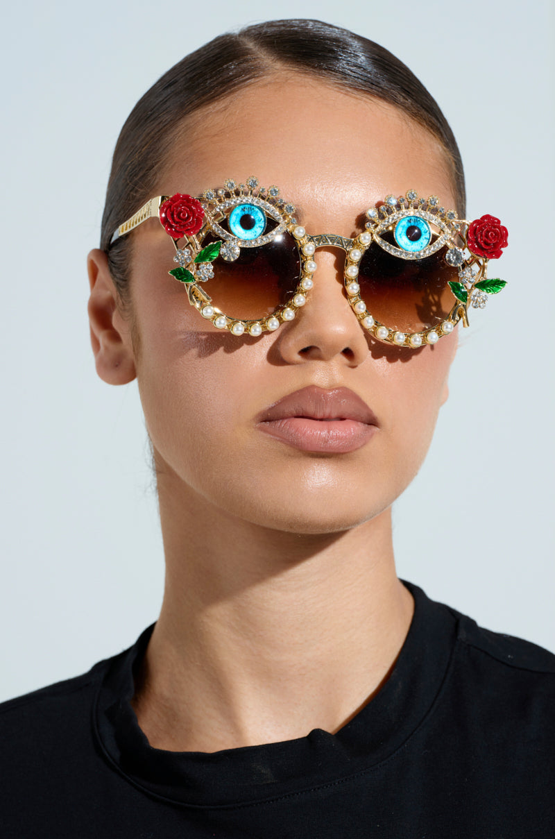 Wearing OFF THE RUNWAY SUNGLASSES featuring ornate gold frames, designs of blue eyes, red roses, and pearl-like rhinestones. They maintain a neutral expression in a black top against a plain backdrop. 3
