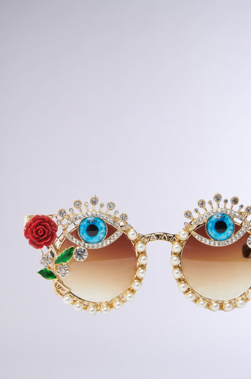 The OFF THE RUNWAY SUNGLASSES feature gold frames with rhinestone accents and eyeglass lenses resembling blue eyes, adorned with a red rose embellishment on the left corner for an elegant touch. 4