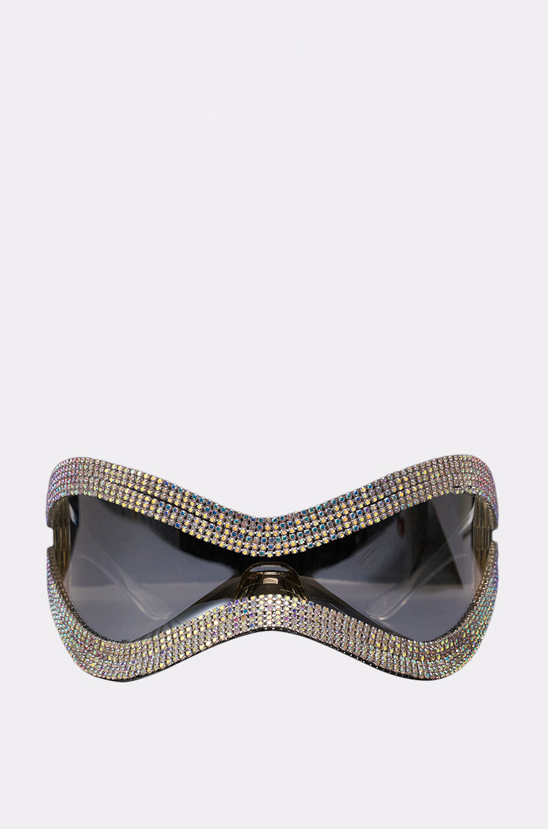 The SHIELD YOU BLING GLASSES are oversized, futuristic shield sunglasses with dark lenses, a curved frame, and fully encrusted multicolored rhinestones—an eye-catching festival accessory that stands out against a plain white background.