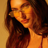 A woman with long brown hair confidently poses in warm, golden light, wearing a colorful striped top and the AKIRA CARTI SUNGLASSES with rectangular yellow-tinted lenses, subtly enhanced by rhinestone embellishments.