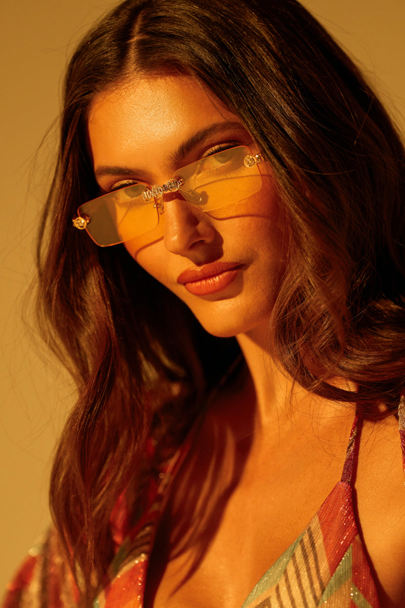 A woman with long brown hair confidently poses in warm, golden light, wearing a colorful striped top and the AKIRA CARTI SUNGLASSES with rectangular yellow-tinted lenses, subtly enhanced by rhinestone embellishments.
