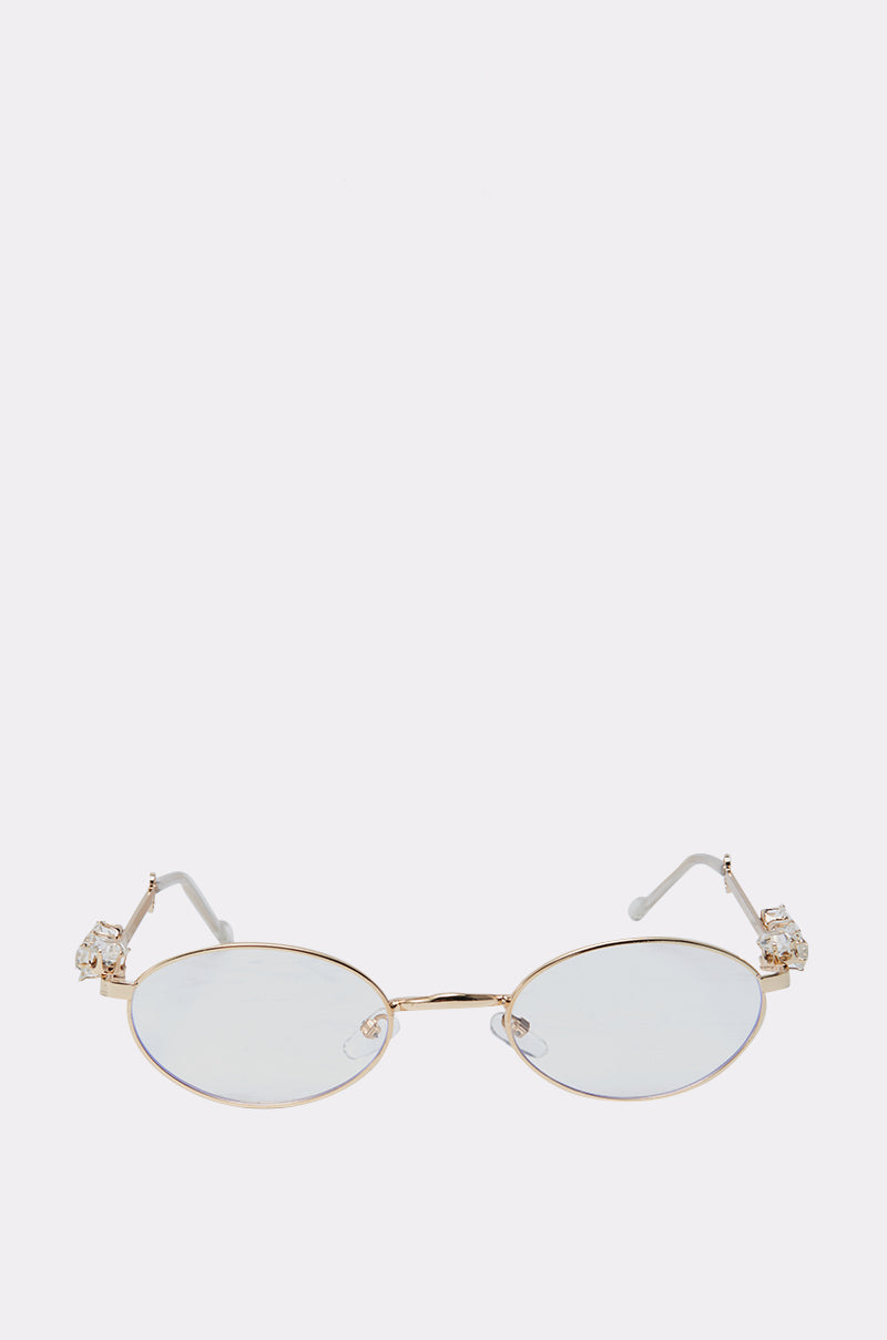 PIERCED VIBES CLEAR GLASSES are gold-rimmed oval eyeglasses with a gold metal frame, clear lenses, and crystal-embellished hinges, displayed on a plain light gray background. 0