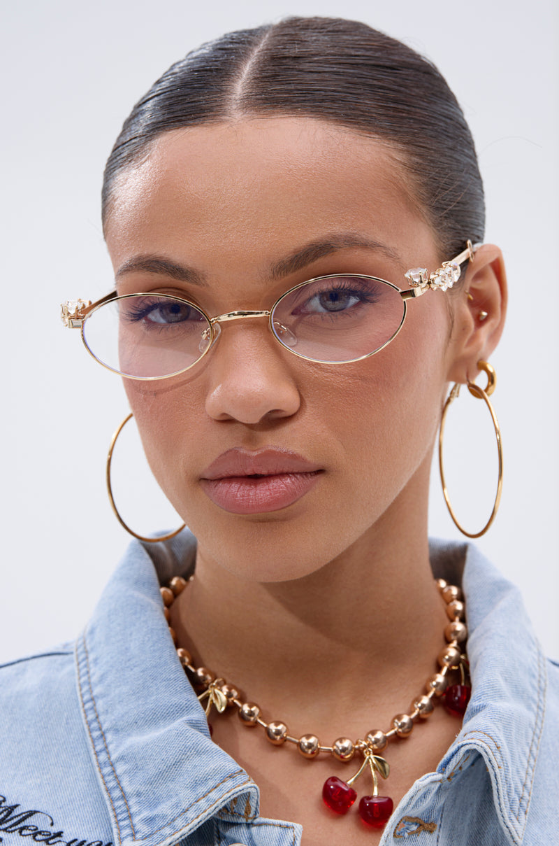 A woman with slicked-back hair wears PIERCED VIBES CLEAR GLASSES with gold frames and decorative details, large gold hoop earrings, a gold beaded cherry charm necklace, and a light denim jacket. Her expression is neutral. 1