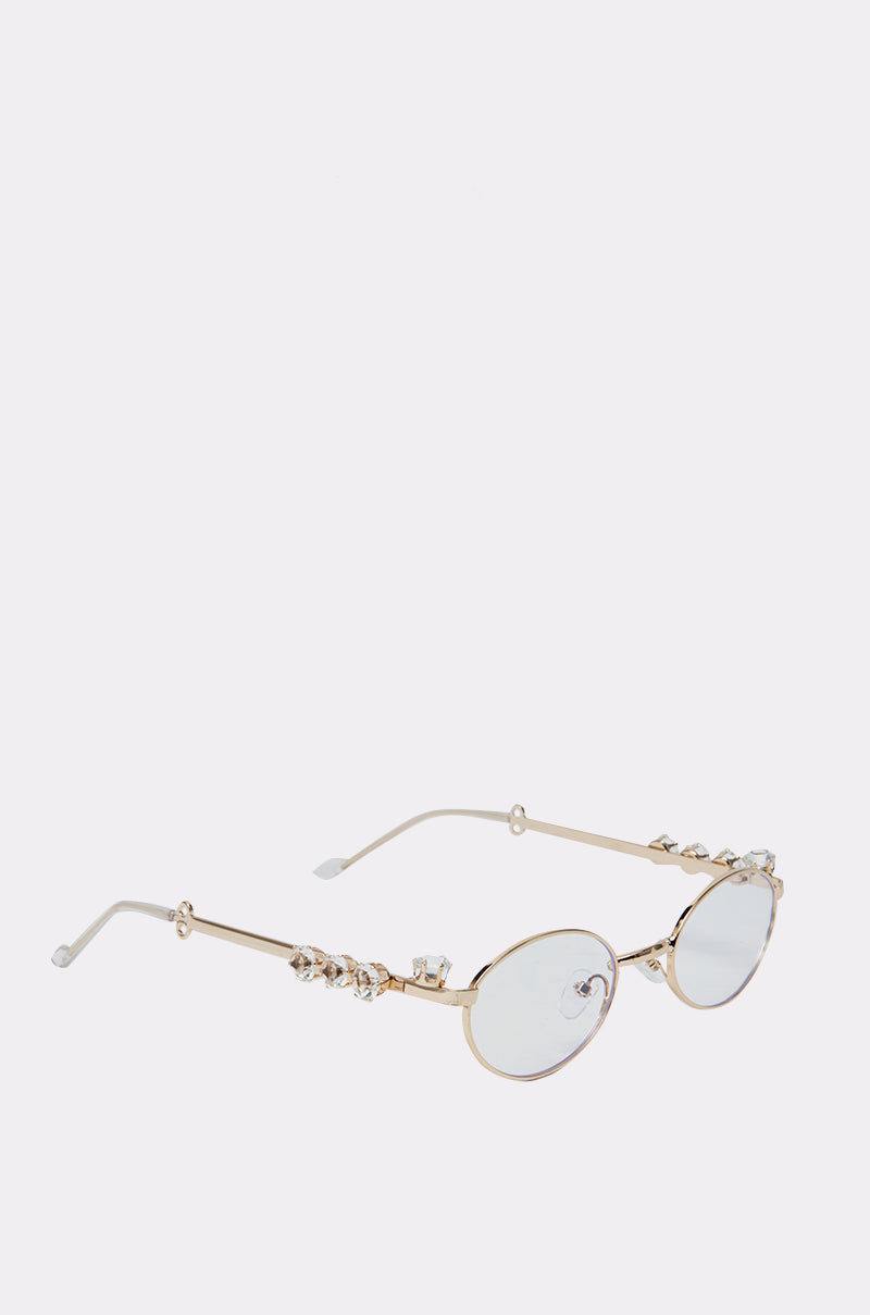 PIERCED VIBES CLEAR GLASSES feature an oval gold-tone metal frame with clear lenses and decorative crystal accents on the temples, shown against a plain white background. 3