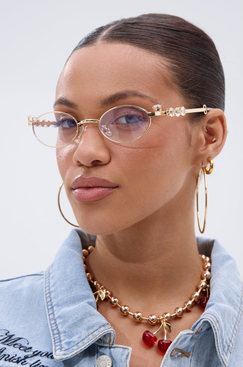 A woman wears PIERCED VIBES CLEAR GLASSES with gold-tone metal frames and decorative jewels, large hoop earrings, a cherry gold necklace, and a light blue denim jacket. Her hair is slicked back and she has a neutral expression. 2
